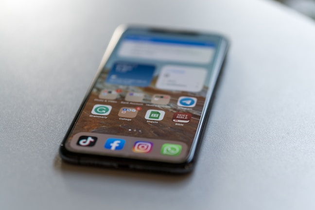 A smartphone is lying on a flat surface, displaying a home screen with various app icons including TikTok, Facebook, Instagram, and WhatsApp. The background is blurred, focusing on the phone.