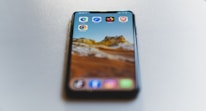 A smartphone is placed on a smooth, light-colored surface. The phone displays a scenic mountain landscape as the wallpaper. Several browser app icons, including Edge, Safari, Chrome, Firefox, and Brave, are visible on the screen.