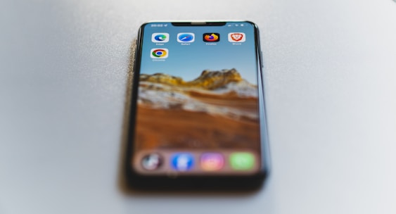 A smartphone is placed on a smooth, light-colored surface. The phone displays a scenic mountain landscape as the wallpaper. Several browser app icons, including Edge, Safari, Chrome, Firefox, and Brave, are visible on the screen.
