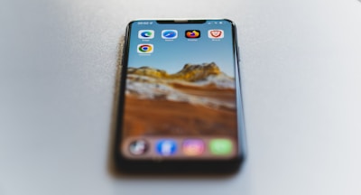 A smartphone is placed on a smooth, light-colored surface. The phone displays a scenic mountain landscape as the wallpaper. Several browser app icons, including Edge, Safari, Chrome, Firefox, and Brave, are visible on the screen.