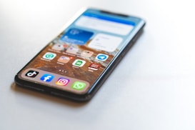 A smartphone placed on a light surface displaying a variety of app icons on the home screen. The phone has a sleek design with an edge-to-edge display. Notable apps include TikTok, Facebook, Instagram, and WhatsApp.