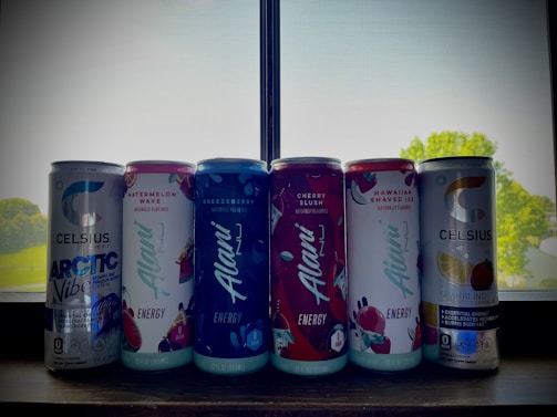 A row of six energy drink cans is placed on a windowsill. The cans include various flavors with vibrant labels, such as Arctic Vibe, Watermelon Wave, Breezberry, Cherry Slush, Hawaiian Shaved Ice, and Sparkling Lemon Lime. The cans are well-lit from behind by natural light streaming in through the window, and there are hints of outdoor greenery visible through the screen.