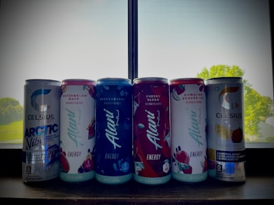A row of six energy drink cans is placed on a windowsill. The cans include various flavors with vibrant labels, such as Arctic Vibe, Watermelon Wave, Breezberry, Cherry Slush, Hawaiian Shaved Ice, and Sparkling Lemon Lime. The cans are well-lit from behind by natural light streaming in through the window, and there are hints of outdoor greenery visible through the screen.
