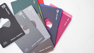 A collection of color sample cards, each labeled with a number and a color name. The cards have different shades such as black, gray, green, blue, and red. They are fanned out on a light surface, and each card has a cutout design.