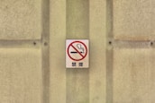 A no smoking sign with a red circle and slash over a cigarette, mounted on a textured beige wall with screws.