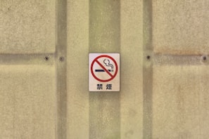 A no smoking sign with a red circle and slash over a cigarette, mounted on a textured beige wall with screws.