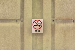 A no smoking sign with a red circle and slash over a cigarette, mounted on a textured beige wall with screws.