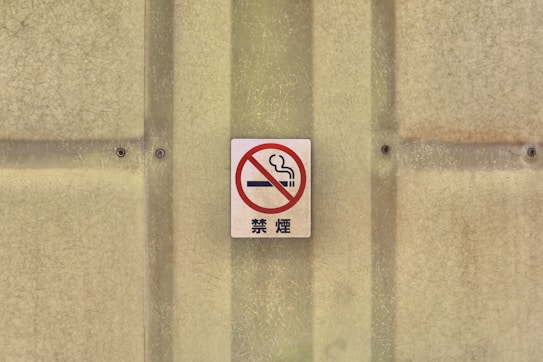 A no smoking sign with a red circle and slash over a cigarette, mounted on a textured beige wall with screws.