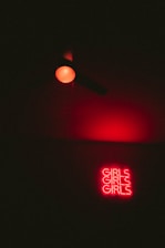 A red neon sign with the word 'GIRLS' repeated three times is illuminated against a dark background. Above it, there is a single red light bulb casting a red glow.