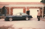 A vintage car is parked in front of a garage with a red brick facade and white doors. A person wearing a beige sweater and dark pants walks beside the car. The area has a rustic, slightly worn look with a concrete driveway.
