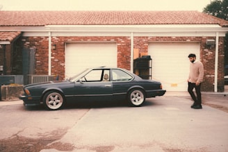 A vintage car is parked in front of a garage with a red brick facade and white doors. A person wearing a beige sweater and dark pants walks beside the car. The area has a rustic, slightly worn look with a concrete driveway.