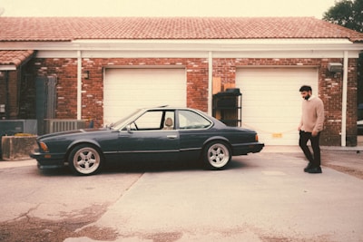 A vintage car is parked in front of a garage with a red brick facade and white doors. A person wearing a beige sweater and dark pants walks beside the car. The area has a rustic, slightly worn look with a concrete driveway.