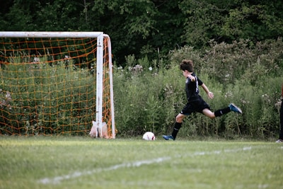 A young player in a black soccer uniform is in motion, preparing to kick a soccer ball towards a goalpost. The goalpost has an orange net and is placed against a backdrop of lush greenery.