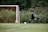 A young player in a black soccer uniform is in motion, preparing to kick a soccer ball towards a goalpost. The goalpost has an orange net and is placed against a backdrop of lush greenery.