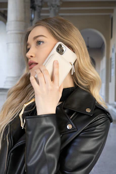 A person with long blonde hair is holding a smartphone to their ear, dressed in a black leather jacket. The background features an arched hallway with soft lighting.