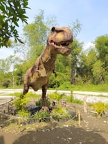 A large and realistic-looking dinosaur sculpture stands in an outdoor setting surrounded by lush greenery and small plants. The dinosaur resembles a Tyrannosaurus Rex, with its mouth open displaying sharp teeth. The scene is set on a sunny day with clear skies.