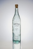 A tall, clear glass bottle with a cork stopper. The bottle has bubbles trapped within the glass and a label featuring the brand name 'Porfidio'. The text indicates it's made from 100% blue agave, cold-pressed, and triple-distilled.