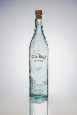 A tall, clear glass bottle with a cork stopper. The bottle has bubbles trapped within the glass and a label featuring the brand name 'Porfidio'. The text indicates it's made from 100% blue agave, cold-pressed, and triple-distilled.
