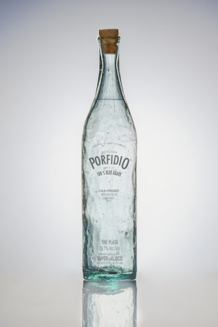 A tall, clear glass bottle with a cork stopper. The bottle has bubbles trapped within the glass and a label featuring the brand name 'Porfidio'. The text indicates it's made from 100% blue agave, cold-pressed, and triple-distilled.