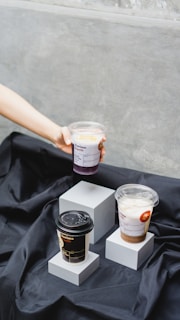 Three beverages are displayed on small white pedestals covered with a black fabric. Two of the drinks are in clear plastic cups with lids, one held by a hand extending from the left, and the other resting on a pedestal. The third beverage is in a black takeaway coffee cup with a lid.
