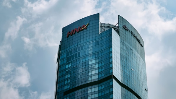A modern skyscraper with a curved glass facade, prominently displaying the HNA logo at the top. The building rises against a backdrop of a cloudy sky, showcasing a blend of architectural innovation and corporate branding.