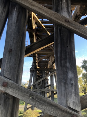 A wooden bridge structure with large timber beams and cross-bracing. The wood appears weathered, with sunlight casting shadows and creating a pattern on the beams. The sky is partly visible with some trees in the background.