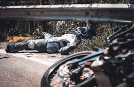 A person wearing motorcycle gear and a helmet is lying on the ground next to a metal guardrail. A motorcycle is seen in the foreground, slightly out of focus. The environment has vegetation and appears to be near a road.