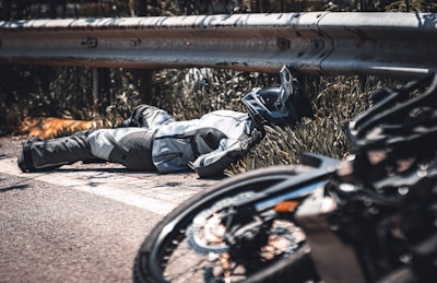 A person wearing motorcycle gear and a helmet is lying on the ground next to a metal guardrail. A motorcycle is seen in the foreground, slightly out of focus. The environment has vegetation and appears to be near a road.