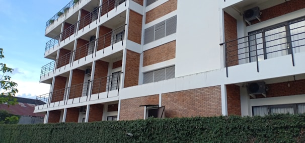 A multi-story building featuring a combination of white and brick exterior with numerous balconies and black railings. Air conditioning units are visible on some balconies. A hedge runs along the base of the building, and a small portion of a roof is seen in the background.