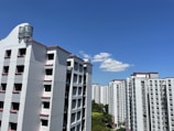 High-rise residential buildings with a white facade and pink accents are seen against a clear blue sky. A few fluffy white clouds are present, and green trees are visible at the base of the buildings.