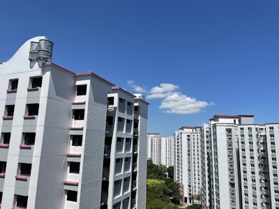 High-rise residential buildings with a white facade and pink accents are seen against a clear blue sky. A few fluffy white clouds are present, and green trees are visible at the base of the buildings.