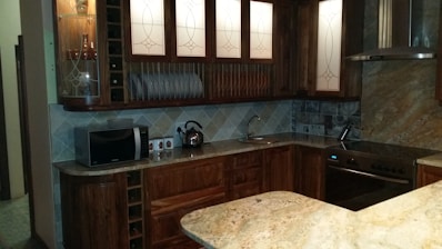 A well-organized kitchen with wooden cabinetry, featuring glass-fronted cupboards and a countertop with a built-in sink. The kitchen is equipped with a microwave, electric kettle, and a stove with an oven. Decorative plates are neatly displayed above the counter, and the backsplash features a pattern of tiled squares.