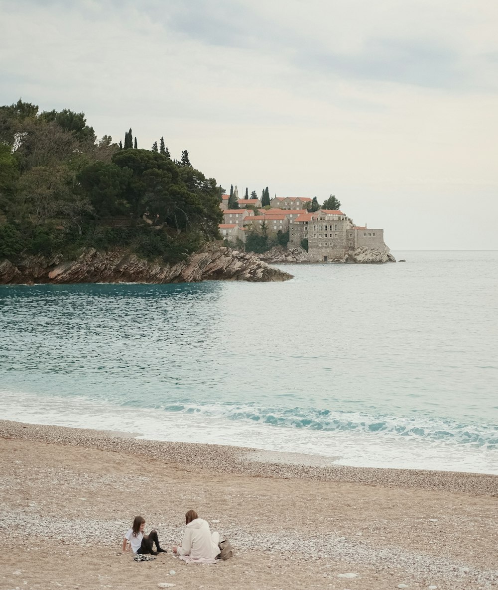 a couple of people sitting on a beach by a body of water