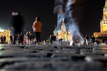 A nighttime scene featuring a cobblestone plaza bustling with people walking, some captured in motion blur. In the background, the illuminated colorful domes of an iconic cathedral rise against the night sky, flanked by other historic buildings with warm lighting.