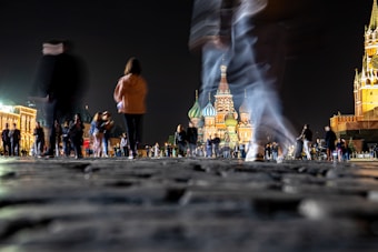 A nighttime scene featuring a cobblestone plaza bustling with people walking, some captured in motion blur. In the background, the illuminated colorful domes of an iconic cathedral rise against the night sky, flanked by other historic buildings with warm lighting.