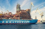 A large ship is docked in a shipyard, undergoing construction or maintenance. The vessel has scaffolding structures built around it, indicating ongoing work. Multiple cranes are visible around the ship, highlighting the industrious atmosphere. The sky is partly cloudy, with a blue hue complementing the ship's color.