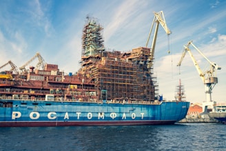 A large ship is docked in a shipyard, undergoing construction or maintenance. The vessel has scaffolding structures built around it, indicating ongoing work. Multiple cranes are visible around the ship, highlighting the industrious atmosphere. The sky is partly cloudy, with a blue hue complementing the ship's color.