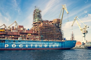 A large ship is docked in a shipyard, undergoing construction or maintenance. The vessel has scaffolding structures built around it, indicating ongoing work. Multiple cranes are visible around the ship, highlighting the industrious atmosphere. The sky is partly cloudy, with a blue hue complementing the ship's color.