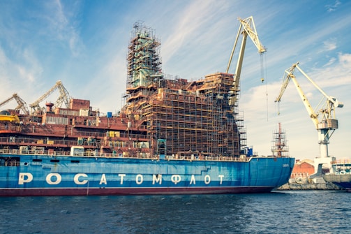 A large ship is docked in a shipyard, undergoing construction or maintenance. The vessel has scaffolding structures built around it, indicating ongoing work. Multiple cranes are visible around the ship, highlighting the industrious atmosphere. The sky is partly cloudy, with a blue hue complementing the ship's color.
