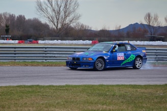 A blue race car is drifting on a track with a slight skid, creating a small plume of smoke from its rear tires. The vehicle has various sponsor stickers, including Liqui Moly, and the number 53 on its side. Behind the car, there is a layering of guardrails, and further back, there are trees with sparse foliage and mountains in the distance under a cloudy sky. The foreground is a grassy area.