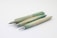 Three wooden mechanical pencils are arranged diagonally on a plain white background. The pencils have a natural greenish tint, with metallic tips and ends. Each pencil displays a unique shade and wood grain pattern, adding a rustic and artisanal appearance.