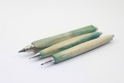 Three wooden mechanical pencils are arranged diagonally on a plain white background. The pencils have a natural greenish tint, with metallic tips and ends. Each pencil displays a unique shade and wood grain pattern, adding a rustic and artisanal appearance.