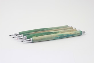 Four mechanical pencils with green and wood-textured barrels are aligned on a light background. The metallic tips and push buttons are visible, with one pencil having a red colored tip.