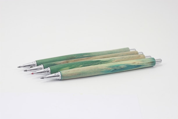 Four mechanical pencils with green and wood-textured barrels are aligned on a light background. The metallic tips and push buttons are visible, with one pencil having a red colored tip.