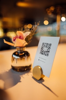 A small, elegant vase with pink and peach flowers is positioned next to a card featuring a QR code. The setup is placed on a table with a warm, ambient background featuring bokeh lighting, creating a cozy and inviting atmosphere.