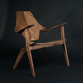 A wooden chair with a modern, angular design is placed against a dark background. The chair appears to have a distinctive, geometric pattern on the backrest and armrests, showcasing intricate craftsmanship in the woodwork.