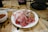 A white plate filled with thin slices of raw meat, including red and pink cuts, is placed on a wooden table. A small bowl containing a brown sauce is positioned to the left of the plate. There are two white chopsticks resting on the table.