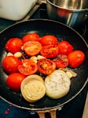 A frying pan contains halved red tomatoes, sliced onions, and garlic cloves being cooked on a stove. The vegetables appear to be slightly charred, suggesting they are being sautéed or grilled. There is a stainless steel pot visible in the background.