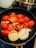 A frying pan contains halved red tomatoes, sliced onions, and garlic cloves being cooked on a stove. The vegetables appear to be slightly charred, suggesting they are being sautéed or grilled. There is a stainless steel pot visible in the background.