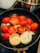 A frying pan contains halved red tomatoes, sliced onions, and garlic cloves being cooked on a stove. The vegetables appear to be slightly charred, suggesting they are being sautéed or grilled. There is a stainless steel pot visible in the background.